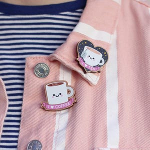 May include: Two enamel pins featuring cartoon mugs with smiling faces. The coffee mug pin has a pink banner with the text "I love coffee". The tea mug pin has a pink banner with the text "I love tea" and a heart shape with stars and moons.