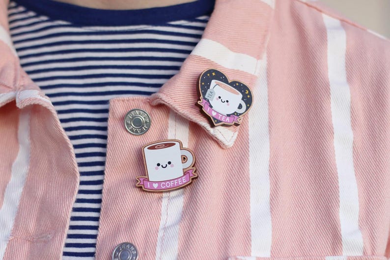 May include: Two enamel pins featuring cartoon mugs with smiling faces. The coffee mug pin has a pink banner with the text "I love coffee". The tea mug pin has a pink banner with the text "I love tea" and a heart shape with stars and moons.