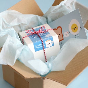 May include: A white box with red and white twine tied around it. The box has a sticker that says "Handmade Pincushion" and a sticker with a cartoon mug and the text "hand made doodle". The box is inside a brown cardboard box with white tissue paper. A card with a cartoon egg and toast is visible in the background.