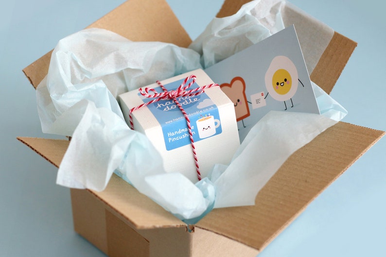 May include: A white box with red and white twine tied around it. The box has a sticker that says "Handmade Pincushion" and a sticker with a cartoon mug and the text "hand made doodle". The box is inside a brown cardboard box with white tissue paper. A card with a cartoon egg and toast is visible in the background.