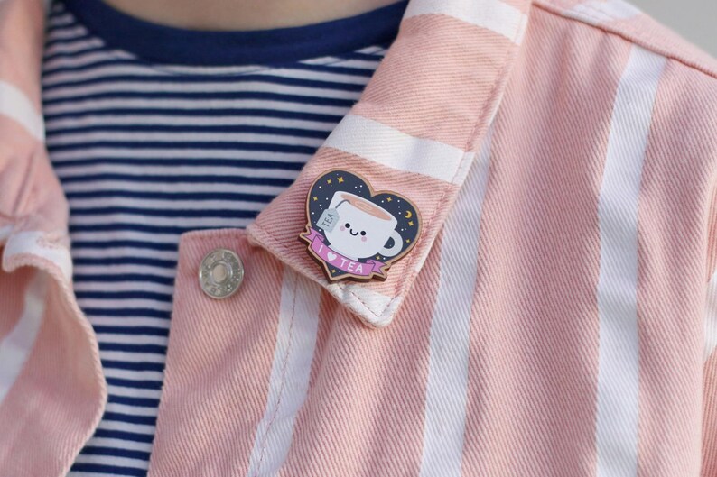 May include: A pink and white striped denim jacket with a wooden pin featuring a cartoon teacup with a tea bag, stars, and a crescent moon. The pin says "I love tea".
