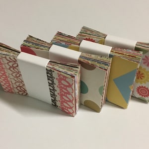 Peut inclure: Quatre piles de cartes en papier &agrave; motifs color&eacute;s, chaque pile est envelopp&eacute;e de papier blanc et attach&eacute;e avec un ruban blanc.