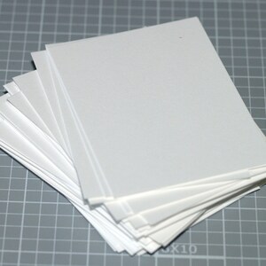 May include: A stack of white, rectangular paper sheets, slightly fanned out, resting on a gray grid surface. The paper appears to be smooth and clean, with a slight sheen. The edges of the paper are crisp and well-defined.