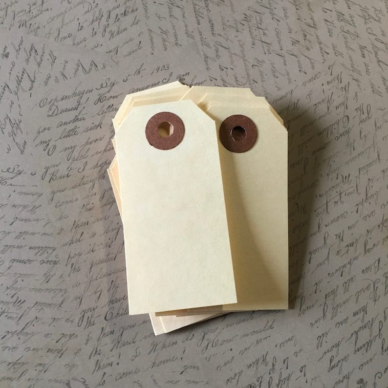 Op de afbeelding: Een stapel blanco, cr&egrave;mekleurige cadeau-etiketten met bruine metalen oogjes. De etiketten zijn perfect om een ​​persoonlijke touch aan geschenken toe te voegen.