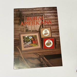 May include: A book cover with the title "Rural Americana" by Jim Anderson. The cover features three framed cross-stitch patterns, two with red and white color schemes and one with green and brown colors. The background is a rustic wooden wall.