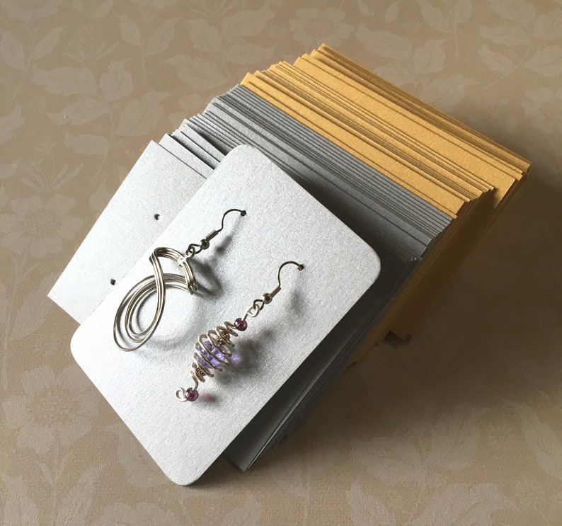May include: Two pairs of handmade earrings displayed on silver earring display cards. One pair features a wire infinity symbol design, and the other has a spiral design with purple beads. A stack of gold and silver cards are in the background.