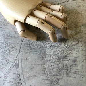 Pu&ograve; includere: Un modello di mano in legno appoggiato su una mappa del mondo vintage. La mappa mostra i continenti di Africa, Asia e Australia. La mano &egrave; in legno marrone chiaro e ha cinque dita.
