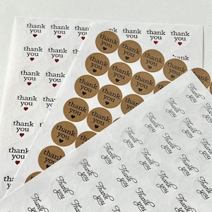 May include: Two sheets of round thank you stickers. One sheet has brown kraft paper stickers with black text and a small heart. The other sheet has clear stickers with black cursive text. Both sheets have the text "Thank You".