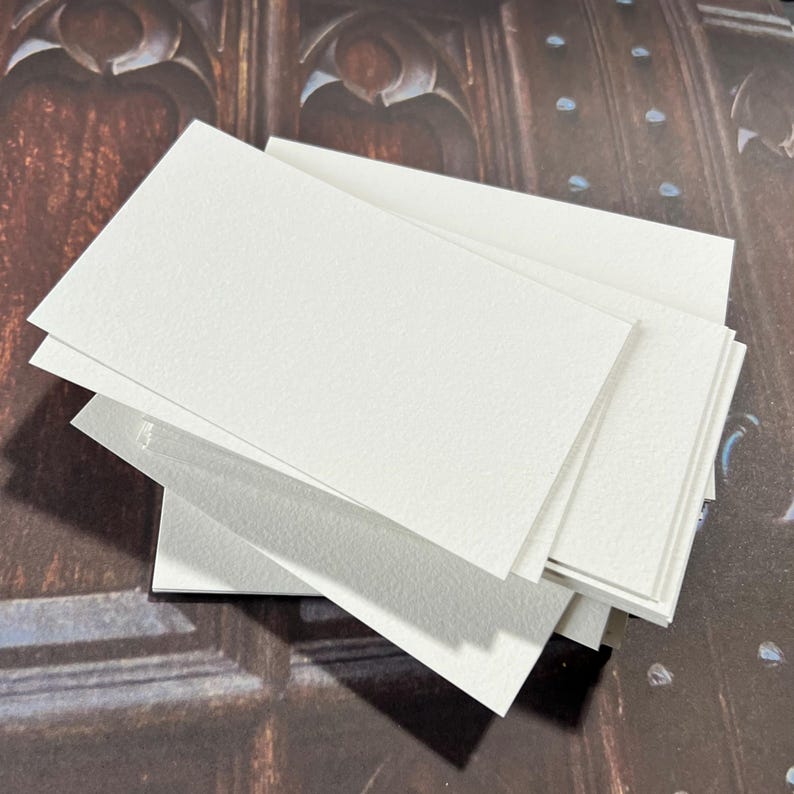 May include: A stack of blank, rectangular, off-white cards. The cards have a slightly textured surface and are arranged in a fan shape, with some cards overlapping others. The background is a blurred image of a dark wooden surface.