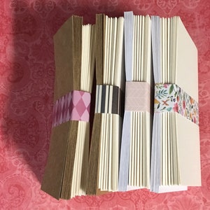 May include: Four stacks of blank note cards with decorative washi tape. The stacks are arranged from left to right with brown, white, white, and white paper. The washi tape is pink and white, black and white, pink and white, and floral.
