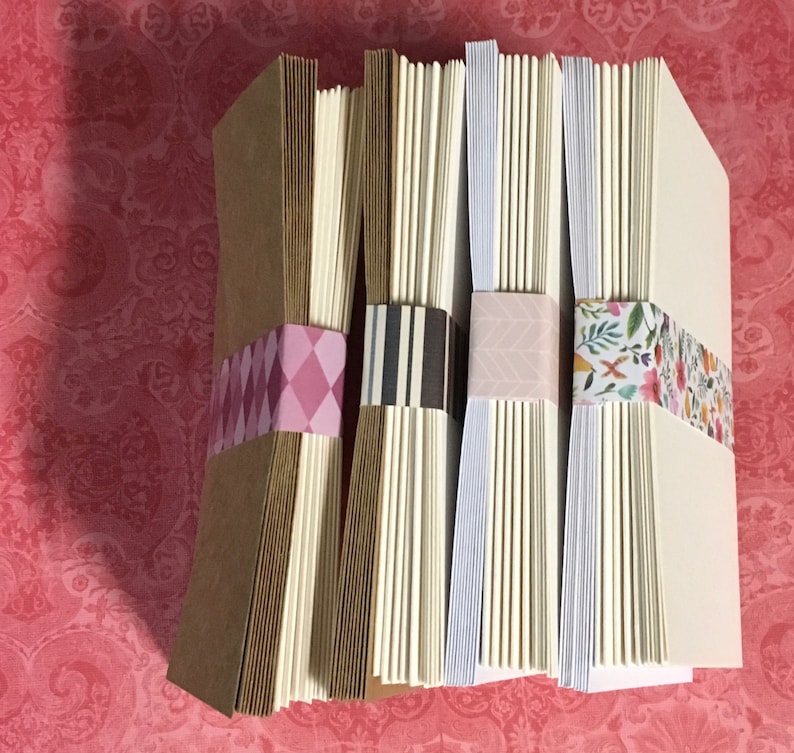 May include: Four stacks of blank note cards with decorative washi tape. The stacks are arranged from left to right with brown, white, white, and white paper. The washi tape is pink and white, black and white, pink and white, and floral.