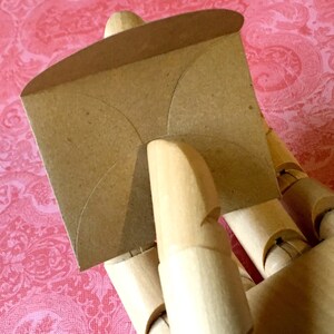 May include: A brown paper envelope with a curved shape, held by a wooden hand.