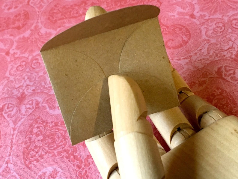 May include: A brown paper envelope with a curved shape, held by a wooden hand.
