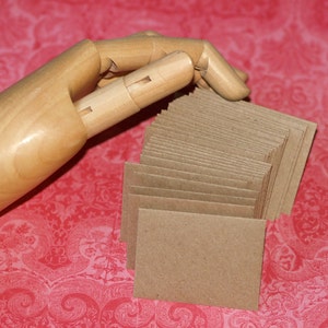 May include: A stack of brown kraft paper envelopes, with a wooden hand holding the top of the stack.