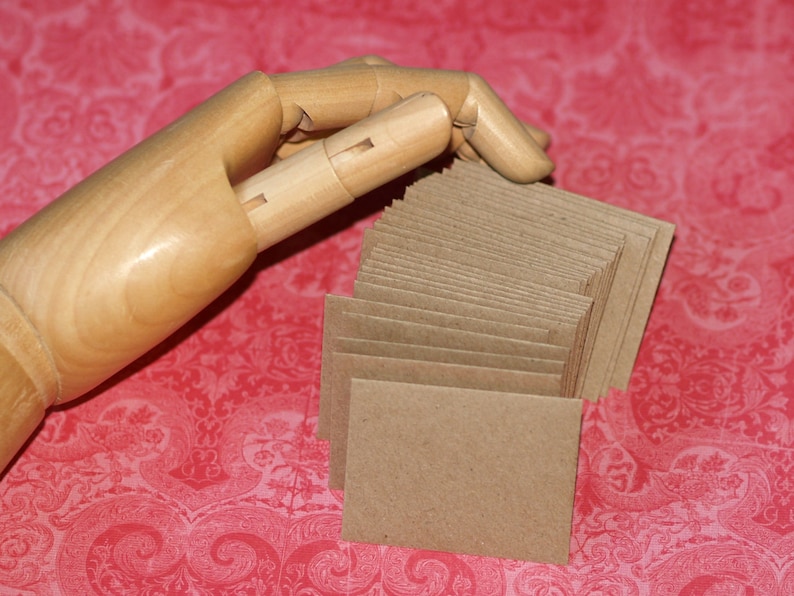 May include: A stack of brown kraft paper envelopes, with a wooden hand holding the top of the stack.
