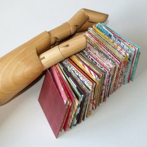 May include: A stack of colorful patterned paper squares bound together to create a small book. The book is held by a wooden hand.