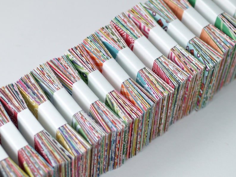 Op de afbeelding: Een stapel kleurrijke vierkante stoffen, elk verpakt in wit papier, gerangschikt in een rij. De stoffen zijn in verschillende patronen en kleuren, waaronder rood, blauw, groen, geel en oranje.