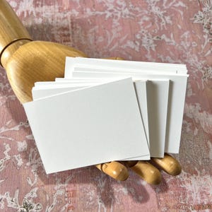 May include: A stack of rectangular white paper cards held by a wooden mannequin hand. The cards are blank, smooth, and opaque, featuring clean, sharp edges. The stack is fanned out slightly, showing multiple layers of thick, white cardstock paper.
