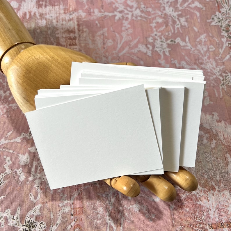May include: A stack of rectangular white paper cards held by a wooden mannequin hand. The cards are blank, smooth, and opaque, featuring clean, sharp edges. The stack is fanned out slightly, showing multiple layers of thick, white cardstock paper.