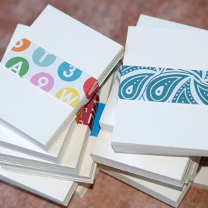 May include: A stack of white note cards with colorful patterned paper wrapped around the edges. One stack has a pattern of letters and numbers, the other has a paisley pattern.