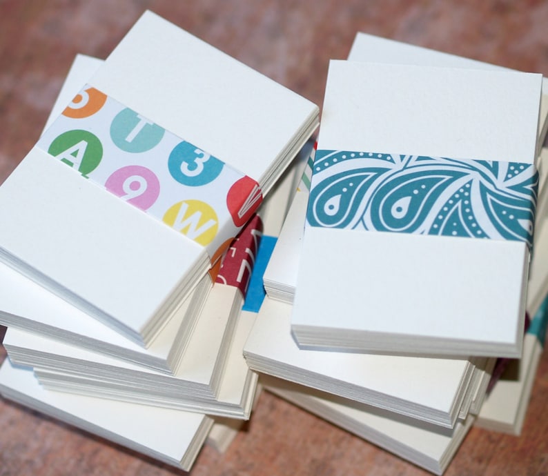 May include: A stack of white note cards with colorful patterned paper wrapped around the edges. One stack has a pattern of letters and numbers, the other has a paisley pattern.