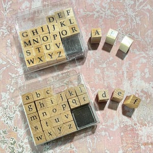 May include: Wooden alphabet stamps in clear plastic cases. The uppercase and lowercase letters are arranged in a grid. Individual letter stamps are scattered around the cases. A black ink pad is included in each case.