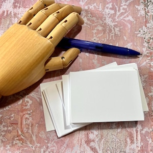 May include: A wooden articulated hand model positioned next to a blue ballpoint pen and a stack of rectangular white paper cards. The items are arranged together, showcasing the natural light wood grain of the hand and the smooth white surface.