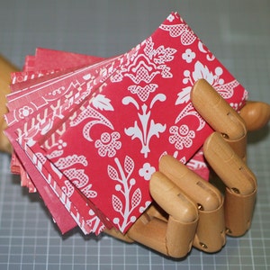 Peut inclure: Une pile d'enveloppes en papier rouge et blanc &agrave; motifs avec un design damass&eacute; blanc. Les enveloppes sont tenues dans une main en bois.