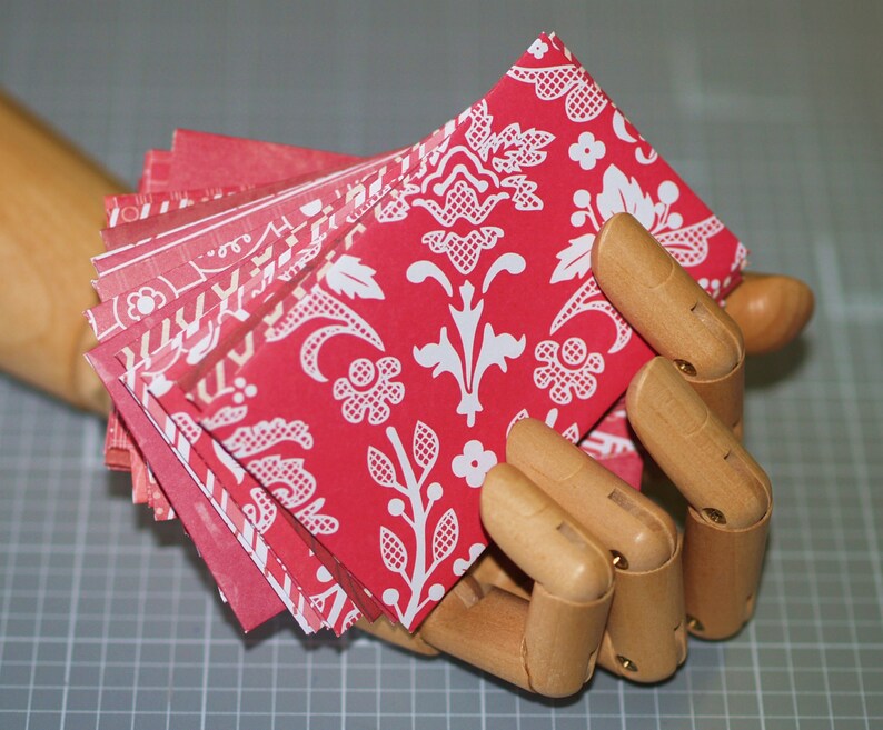 Peut inclure: Une pile d'enveloppes en papier rouge et blanc &agrave; motifs avec un design damass&eacute; blanc. Les enveloppes sont tenues dans une main en bois.