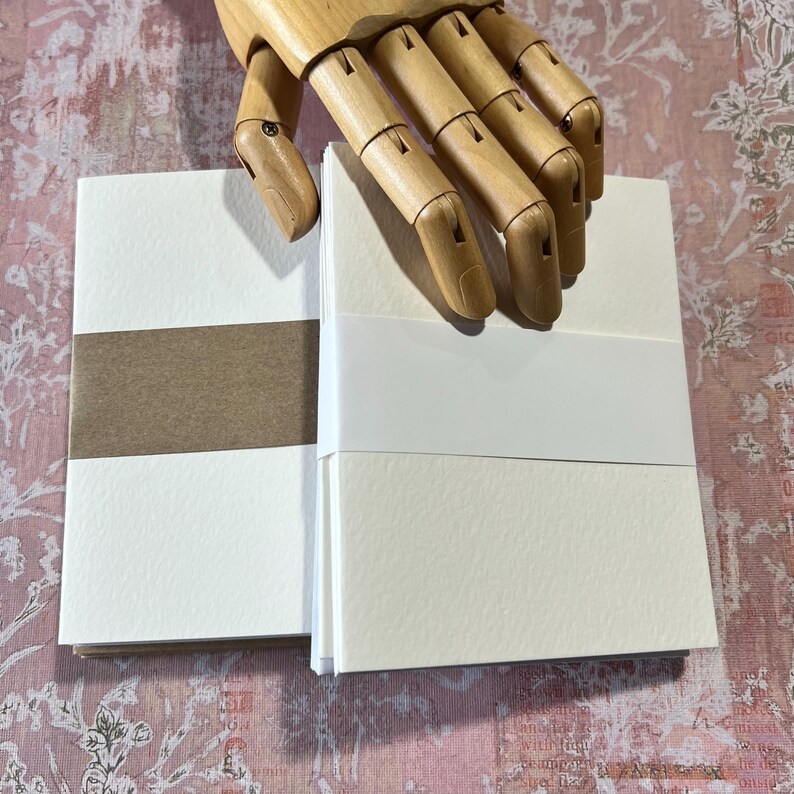May include: Two stacks of blank white note cards with brown paper backing. The cards are arranged vertically and are slightly offset from each other. The cards are on a pink and white floral patterned surface.