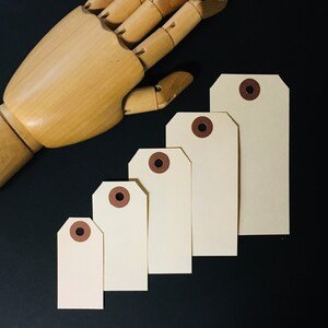 Op de afbeelding: Vijf blanco cadeau-etiketten met bruine oogjes. De etiketten zijn gerangschikt in aflopende volgorde van grootte van links naar rechts. De etiketten bevinden zich op een zwarte achtergrond.