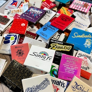 May include: A collection of vintage matchbook covers with various designs and text. The matchbooks feature a variety of businesses and locations, including hotels, restaurants, and motels. Some of the matchbooks have colorful designs, while others are more simple. The matchbooks are a great example of vintage advertising and ephemera.