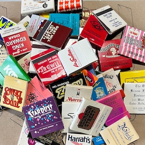 May include: A collection of vintage matchbook covers featuring various businesses and locations. The matchbooks are colorful and feature a variety of designs, including illustrations, text, and logos. Some of the matchbooks are from hotels, restaurants, and casinos. The matchbooks are a great example of vintage advertising and memorabilia.
