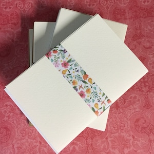 May include: A stack of blank ivory note cards with a floral patterned ribbon wrapped around them. The ribbon has a white background with pink, yellow, and green flowers and leaves.