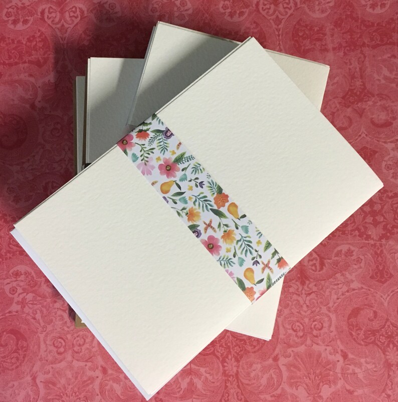 May include: A stack of blank ivory note cards with a floral patterned ribbon wrapped around them. The ribbon has a white background with pink, yellow, and green flowers and leaves.