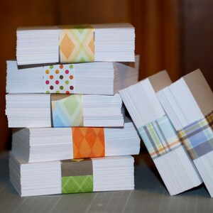 May include: A stack of white card stock paper with colorful patterned washi tape. The stack is made up of five pieces of paper, each with a different patterned tape.