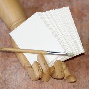 May include: A wooden hand model holding a stack of blank white paper cards and a wooden handled paintbrush.