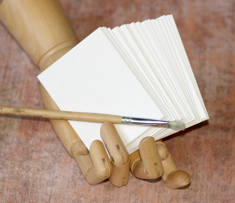 May include: A wooden hand model holding a stack of blank white paper cards and a wooden handled paintbrush.