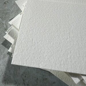 May include: A pile of square, off-white paper sheets with a textured surface. The paper is arranged in a layered formation, with the top sheets in sharp focus. The paper appears to be thick and of high quality.