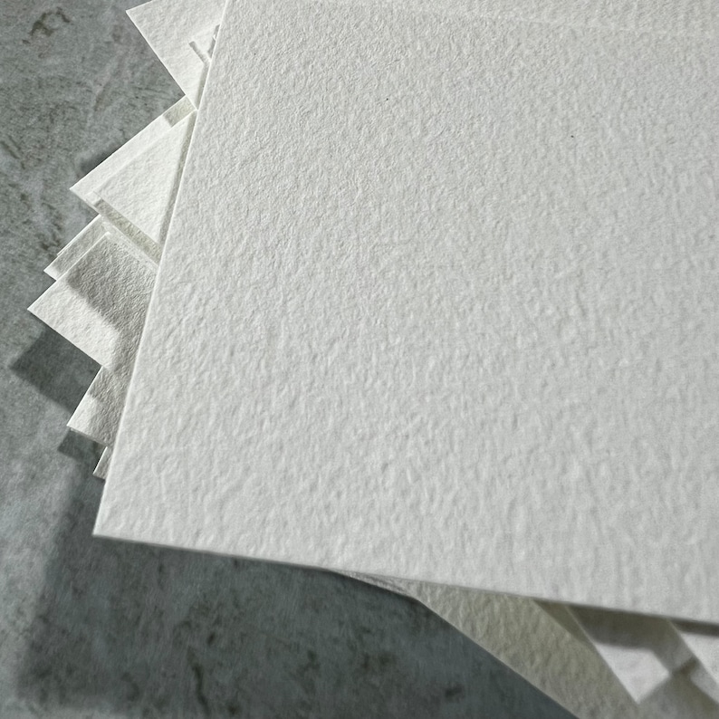 May include: A pile of square, off-white paper sheets with a textured surface. The paper is arranged in a layered formation, with the top sheets in sharp focus. The paper appears to be thick and of high quality.