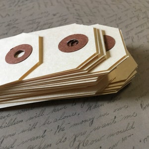 Op de afbeelding: Een stapel beige papieren etiketten met bruine cirkels in het midden. De etiketten zijn dubbelgevouwen en hebben een gegolfde rand.