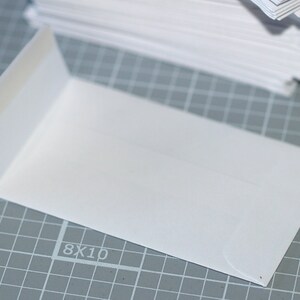 May include: A white envelope with a flap on the left side. The envelope is lying on a gray gridded surface with the number "8X10" printed on it.