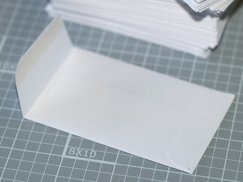 May include: A white envelope with a flap on the left side. The envelope is lying on a gray gridded surface with the number "8X10" printed on it.