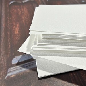 May include: A stack of white, rectangular watercolor paper sheets. The paper has a slightly textured surface and is arranged in a neat pile. The edges of the paper are clean and crisp, suggesting a high-quality art supply.