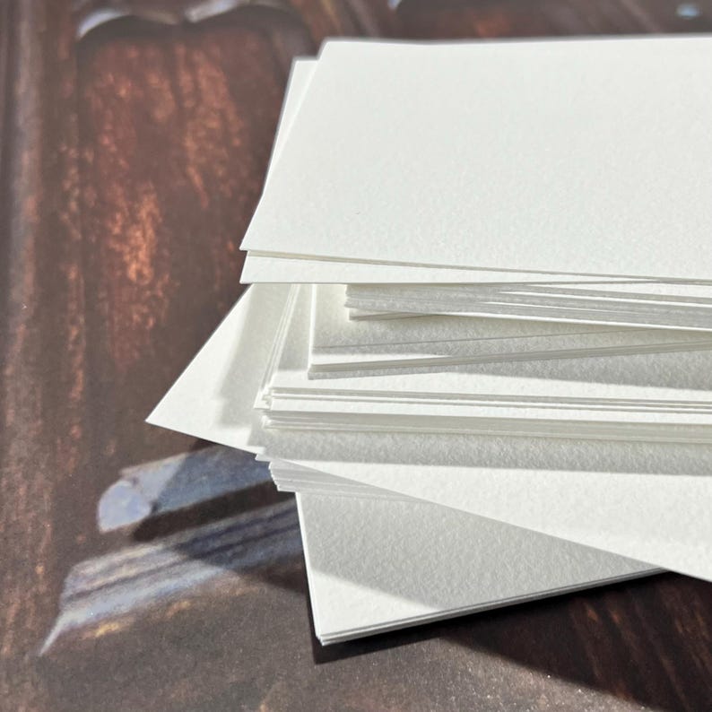 May include: A stack of white, rectangular watercolor paper sheets. The paper has a slightly textured surface and is arranged in a neat pile. The edges of the paper are clean and crisp, suggesting a high-quality art supply.