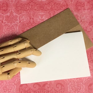 May include: A blank white card with a brown envelope on top of it. The card and envelope are on a pink and white patterned surface.