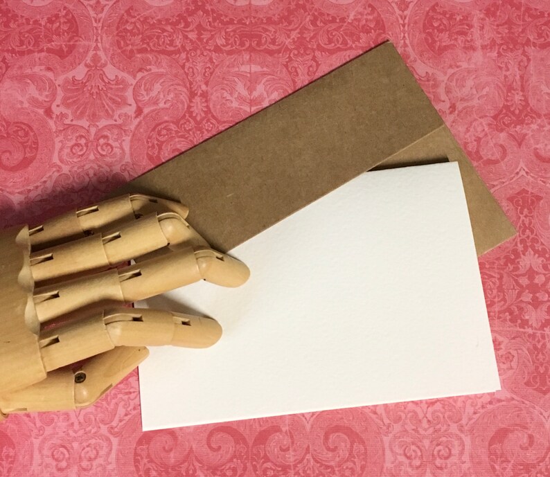 May include: A blank white card with a brown envelope on top of it. The card and envelope are on a pink and white patterned surface.