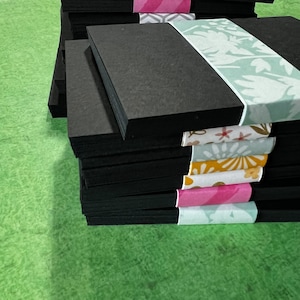 May include: A stack of black cardstock paper with patterned paper accents. The top sheet of cardstock has a light green ribbon with a floral design.