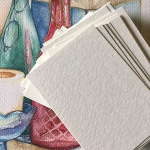 May include: A stack of blank white paper cards with a textured surface. The cards are stacked on top of a watercolour painting of a still life with a green bottle, a red bottle, and books.