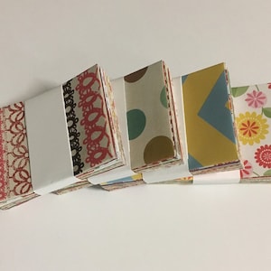 Peut inclure: Quatre piles de carr&eacute;s de papier &agrave; motifs color&eacute;s, chaque pile &eacute;tant envelopp&eacute;e de papier blanc. Les motifs comprennent de la dentelle rouge et blanche, des pois bruns et blancs, des formes g&eacute;om&eacute;triques jaunes et bleues et des motifs floraux roses et jaunes.